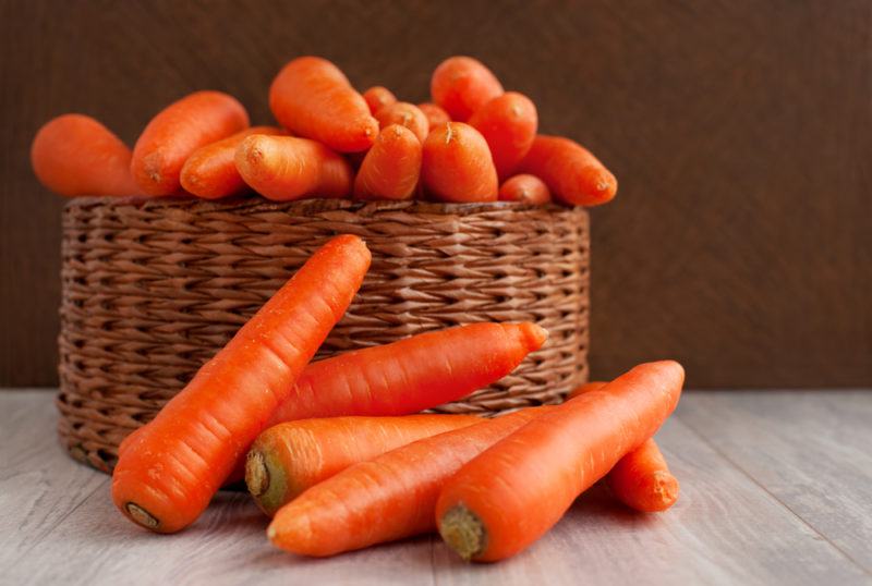 A brown basket containing carrots without their green stalks, with five carrots in front