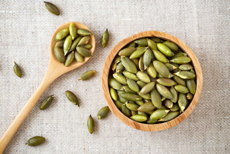 A brown wooden bowl and spoon that contain pumpkin seeds