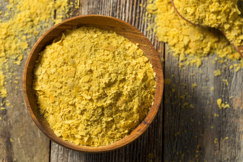 A large wooden bowl of nutritional yeast next to a spoon of the yeast