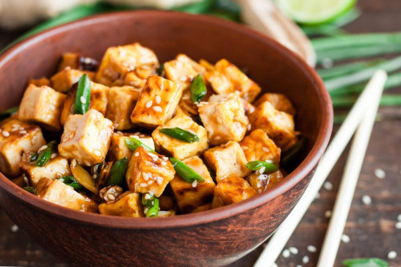 A dark brown bowl with tofu and greens, next to chopsticks