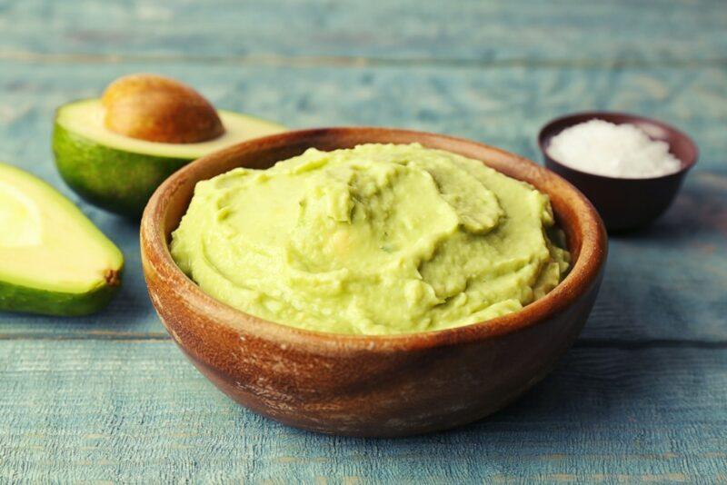 A brown bowl filled with avocado puree, with two avocado halves in the background and a small bowl