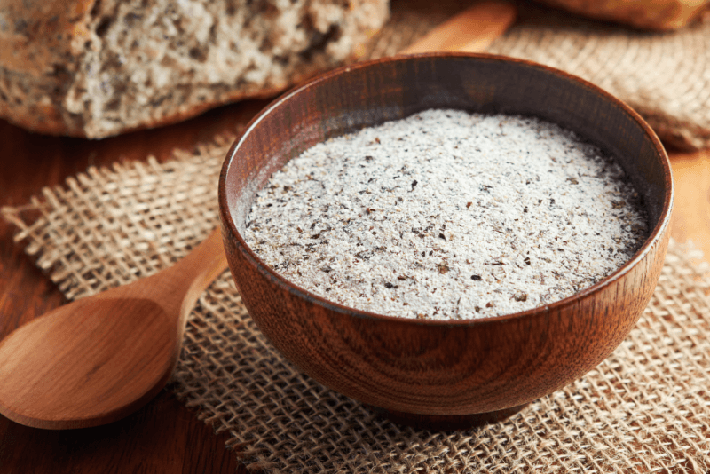 A wooden bowl containing buckwheat flour, next to a wooden spoon, with some bread in the background