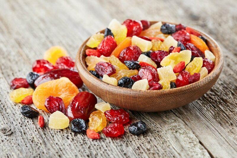 A small brown bowl containing a selection of candied dry fruit, with some more fruit next to it on a wooden table