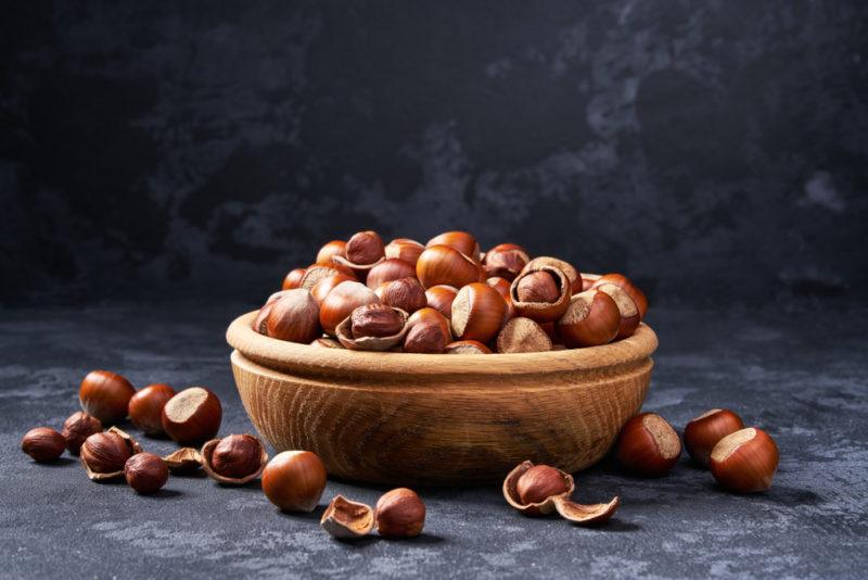 A brown wooden bowl piled high with hazelnuts, with more hazelnuts on the table itself