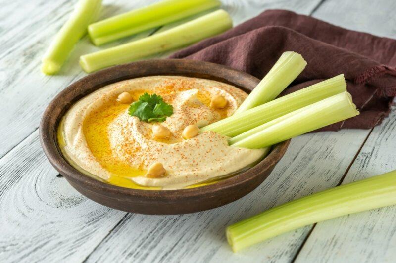 A brown bowl with hummus and celery sticks, with a few more celery sticks on the table