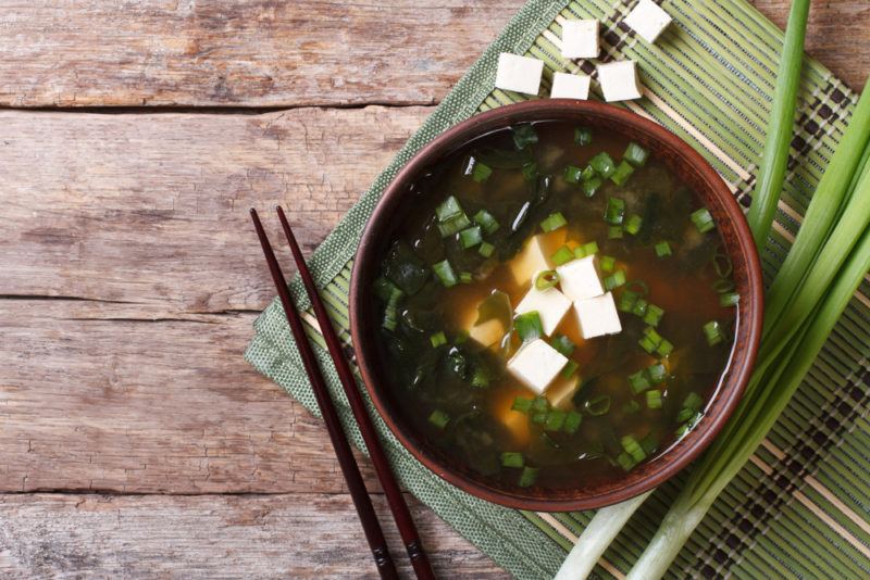 A brown bowl of fresh miso soup with tofu cubes, next to some spring onions, a pair of chopsticks and cubed tofu