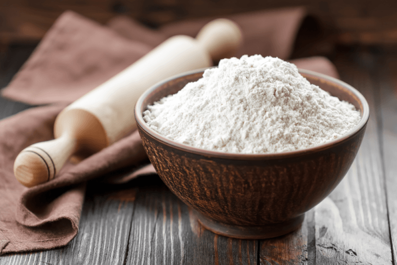 A table that has a brown bowl of self rising flour, next to some cloths and a rolling pin