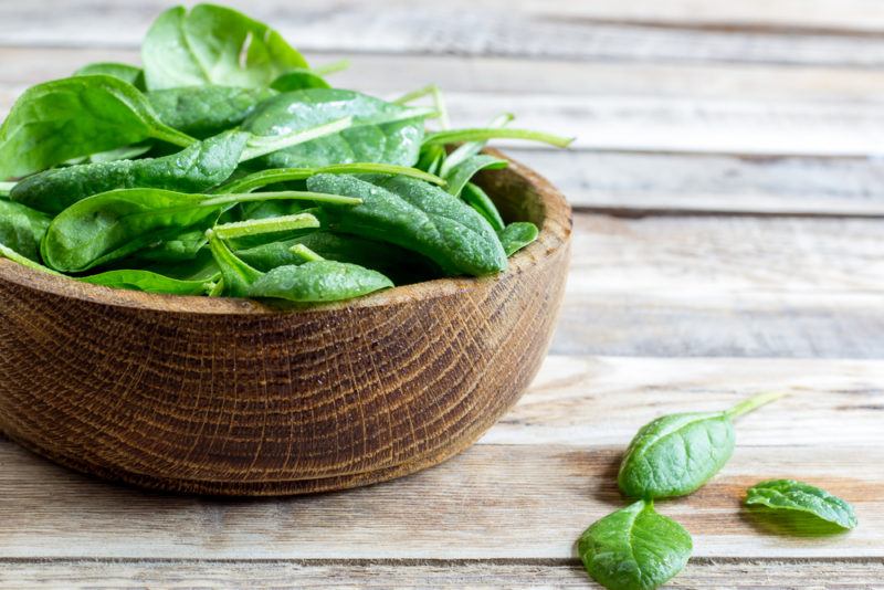 A brown wooden bowl of spinach on a table with a few leaves on the table too