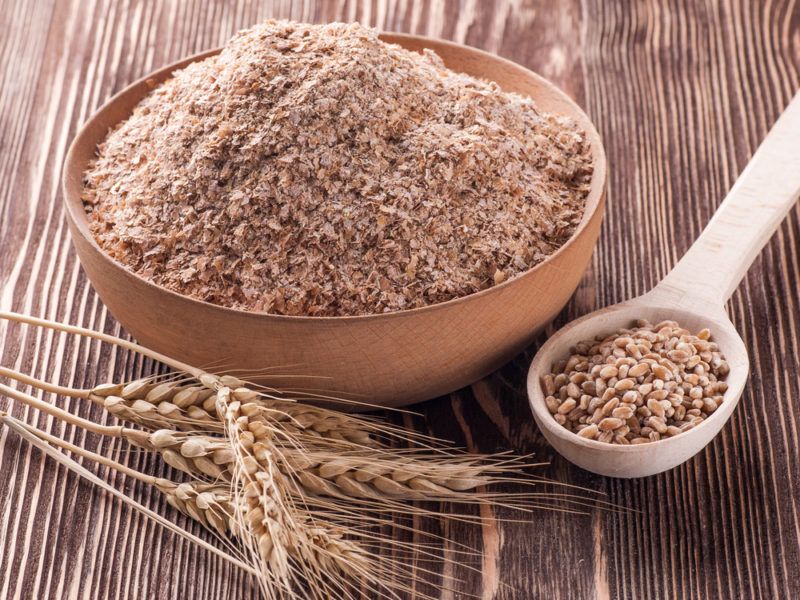 A brown bowl that contains wheat bran next to a spoon of the bran