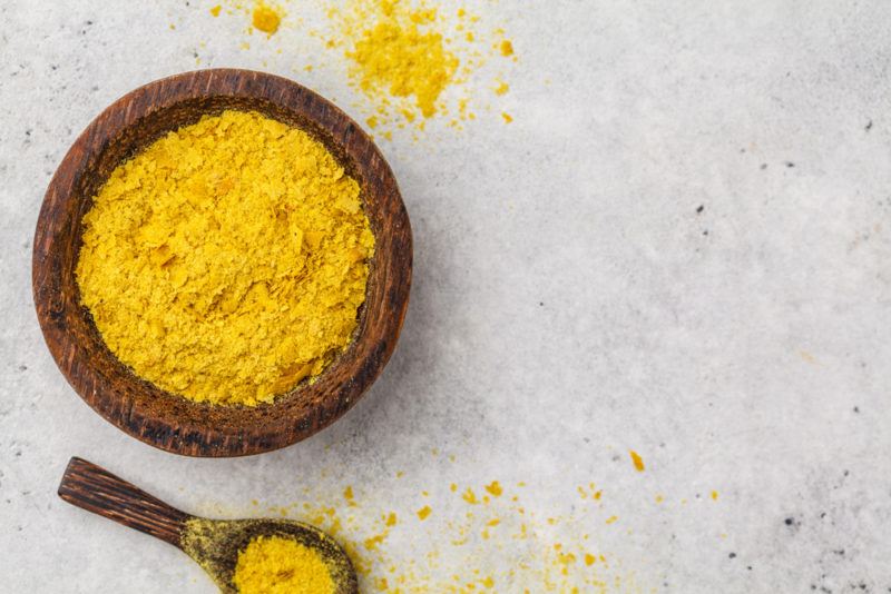 A wooden bowl of nutritional yeast next to a spoon full of the yeast