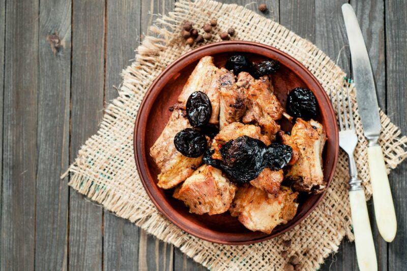 A brown bowl with a meat dish and prunes, next to a knife and fork