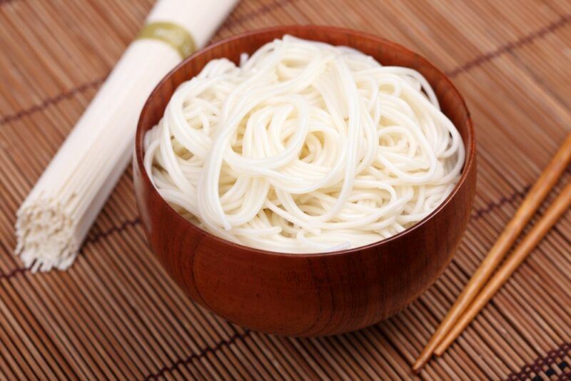 A large brown bowl containing cooked rice noodles, next to some uncooked noodles and chopsticks