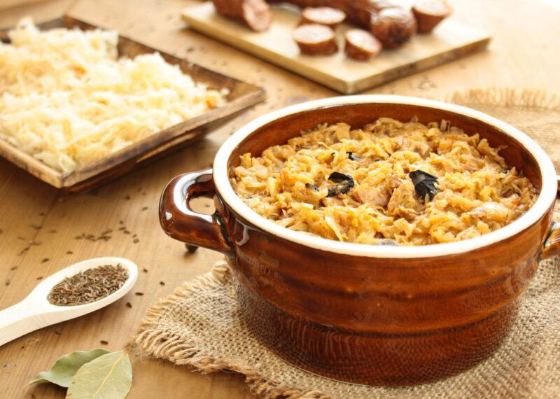 A deep brown dish that contains a soup called bigos, next to a spoon and trays of ingredients