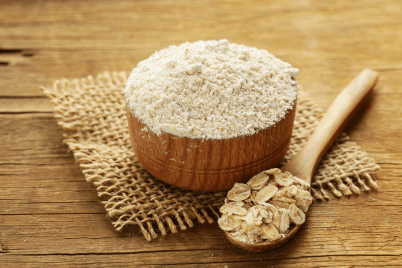 A wooden bowl containing oat flour, next to a wooden spoon of oats. There is a piece of burlap under the bowl, which is on a wooden surface.