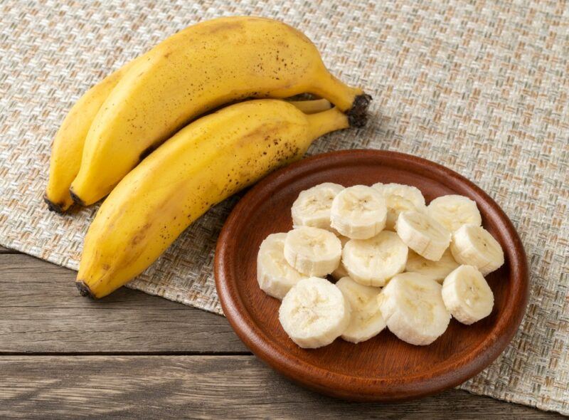 A few fresh bananas, next to a brown plate with sliced bananas