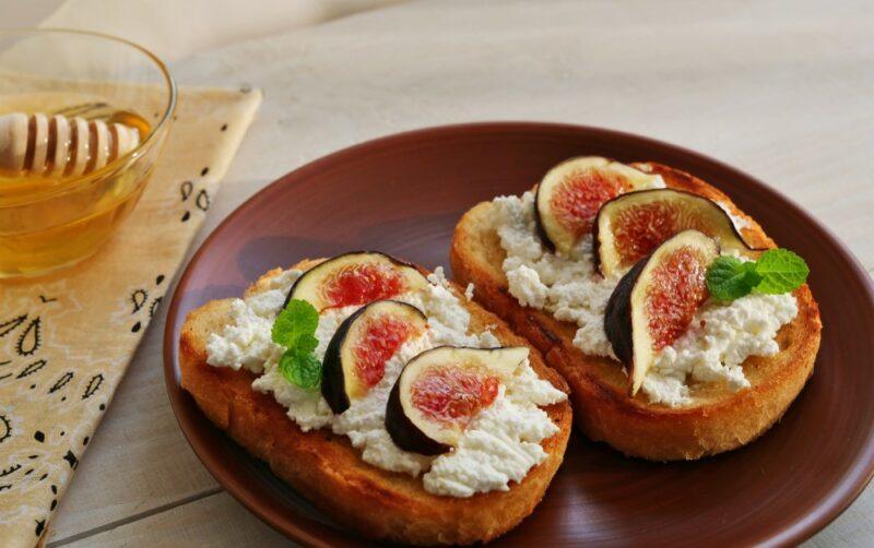 A brown plate with two pieces of bread topped with cottage cheese and sliced figs, with a dish of honey in the background