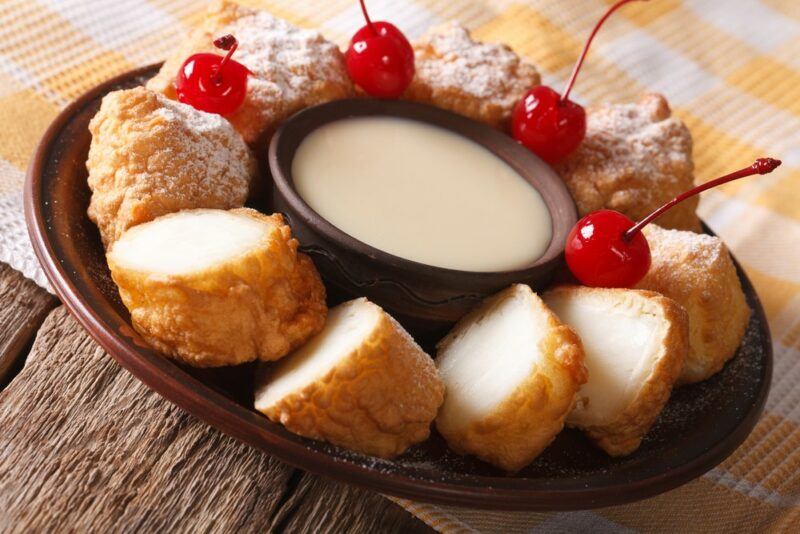 A brown plate with a bowl of dip in the middle and slices of fried milk surrounding it. There are four glace cherries as well