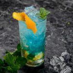 A gray table with a glass of a bullfrog cocktail with ice, garnished with an orange twist