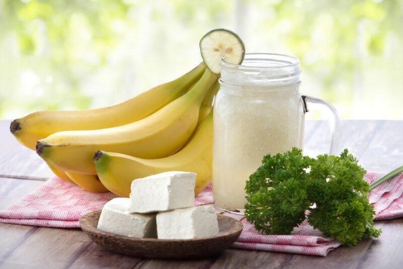 A wooden table with fresh bananas, a smoothie, tofu, and some herbs