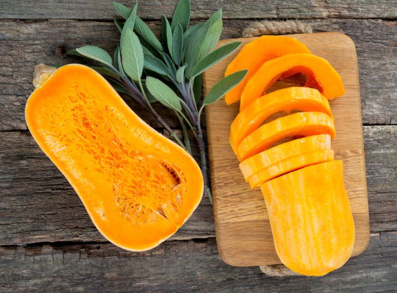 A butternut squash that has been cut in half. One half has been left as-is, while the other has been sliced on a board