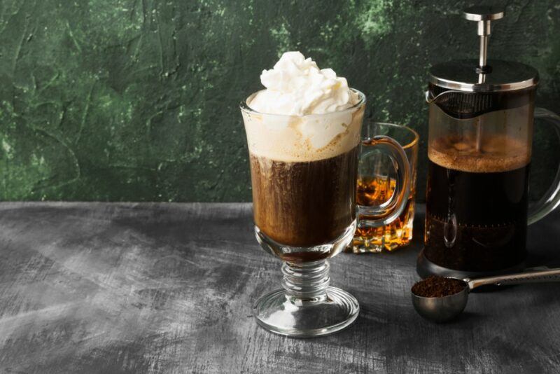 A tall glass containing a caffe di alpi or alpine coffee cocktail with whipped cream on top, next to a glass of whiskey and a coffee plunger