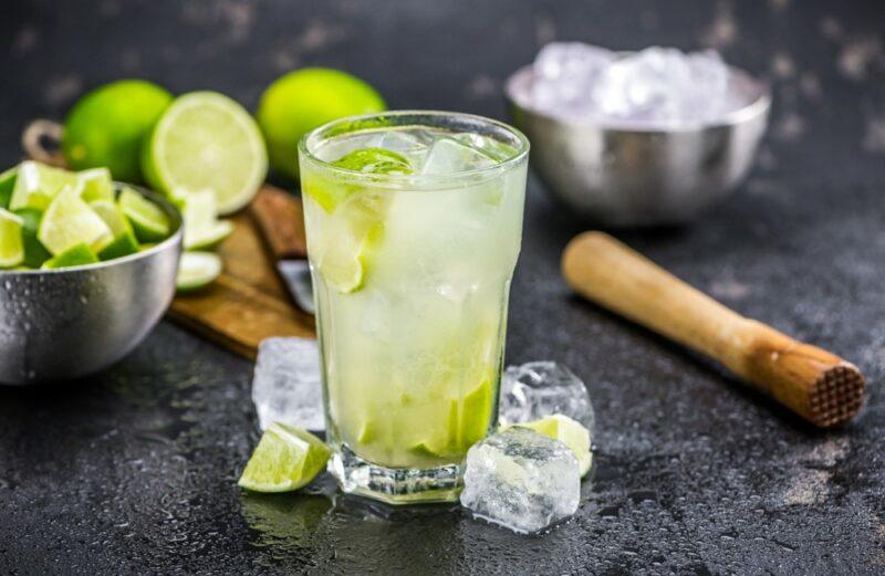 A dark table with a single glass containing a caipirinha cocktail, with limes, ice, and a muddling tool in the background