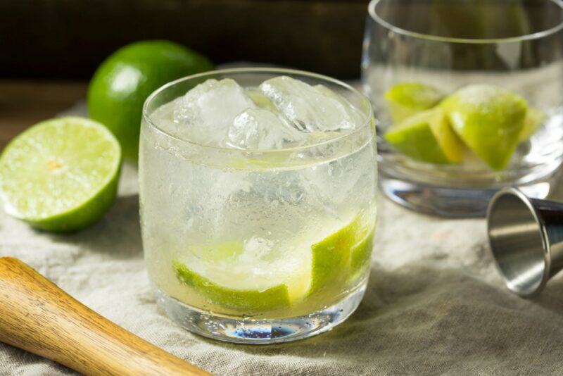 A glass containing a caipirinha cocktail with ice, next to a glass that just contains limes