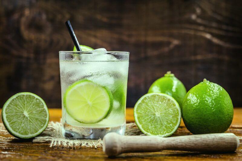 A caipirinha cocktail with ice and lime slices, next to a collection of limes