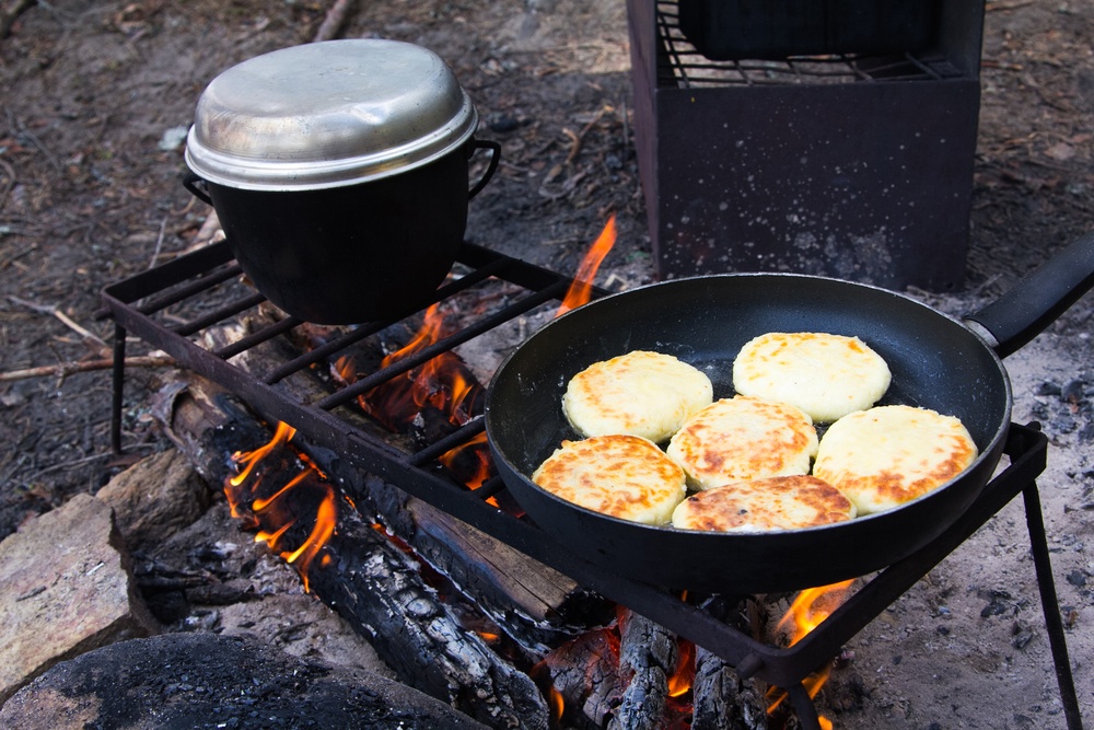 A makeshift grill over a camping fire, with a Dutch oven and pancakes in a frypan