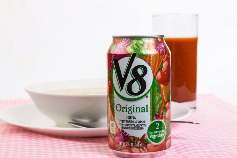 A can of V8 juice in front of a glass and a bowl of soup