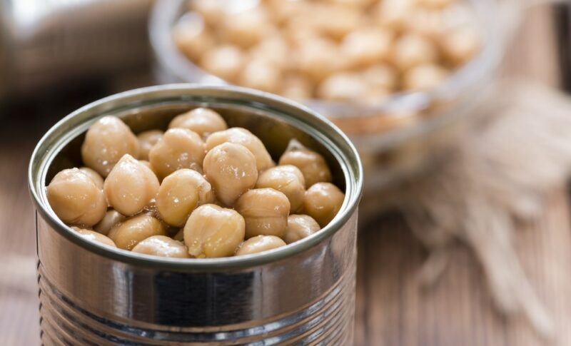 A can of chickpeas in liquid, with a bowl of chickpeas out of focus in the background