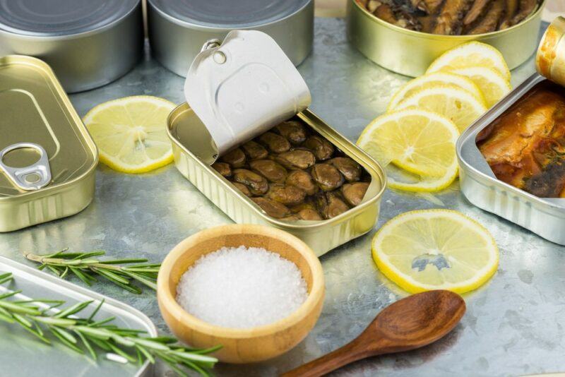 A can of smoked oysters with the lid removed on a table with lemon, herbs, salt, and a spoon