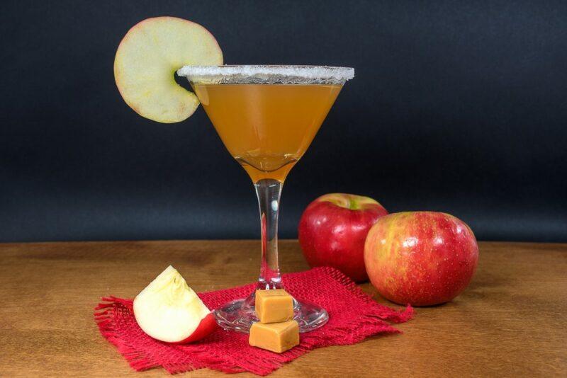 A caramel apple martini that uses an apple round as a garnish, with apples and a few pieces of caramel on the table