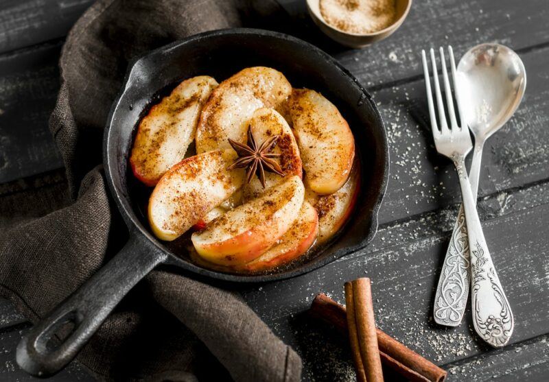 A black cast iron pan with apples and cinnamon, next to a spoon and a fork