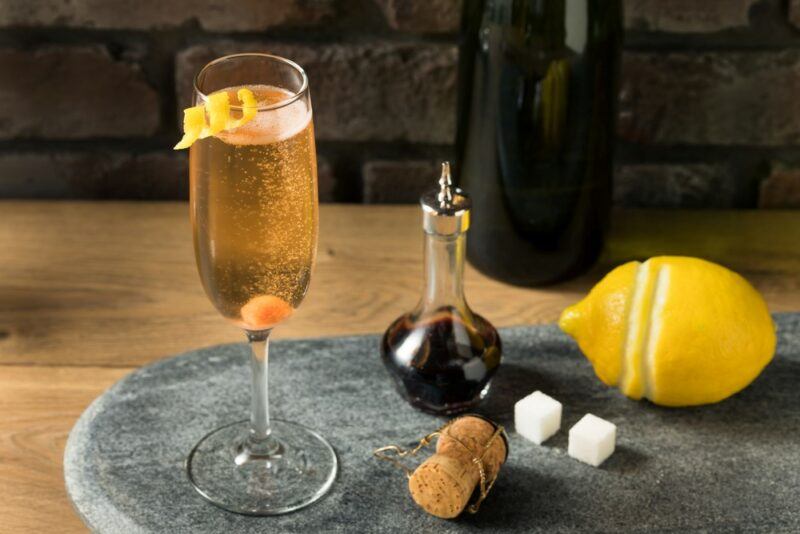 A glass containing a French champagne cocktail, next to some bitters, two sugar cubes, a lemon, and a champagne cork