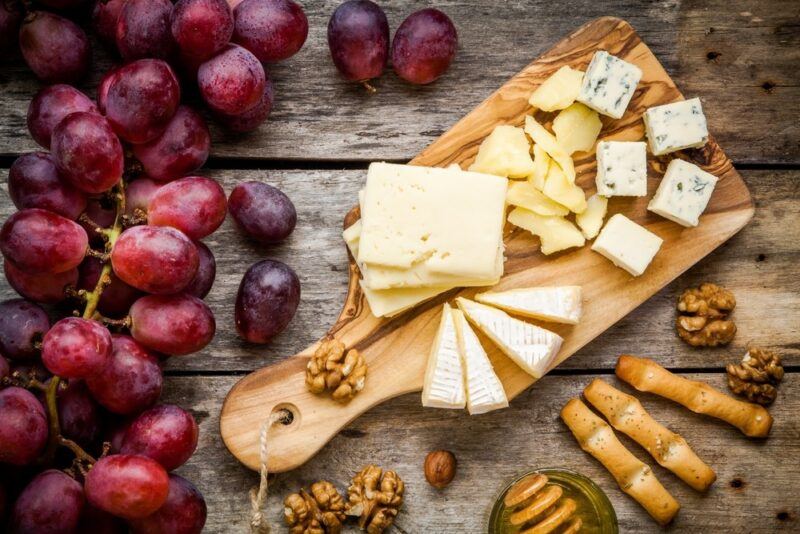 A wooden board with many pieces of cut cheese, next to a pile of fresh red grapes