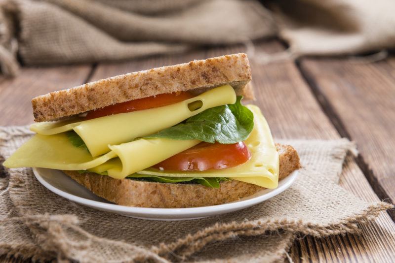 A freshly made sandwich with cheese, spinach and tomato, on a white plate that's on a sack or cloth and a wooden table