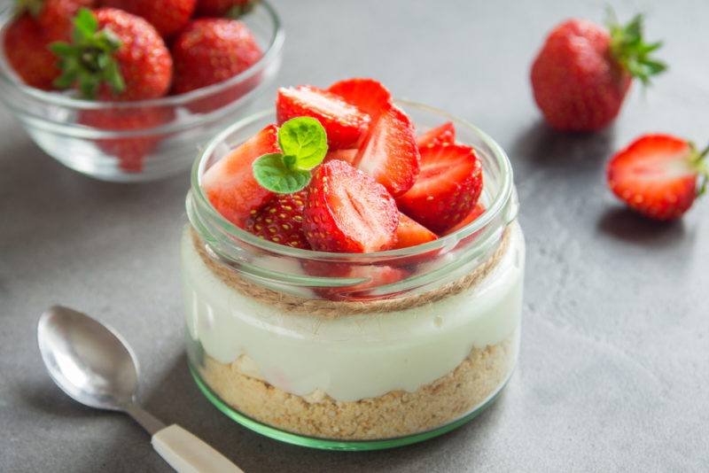 A small jar containing a fresh cheesecake with strawberries on top, and a bowl of strawberries in the background