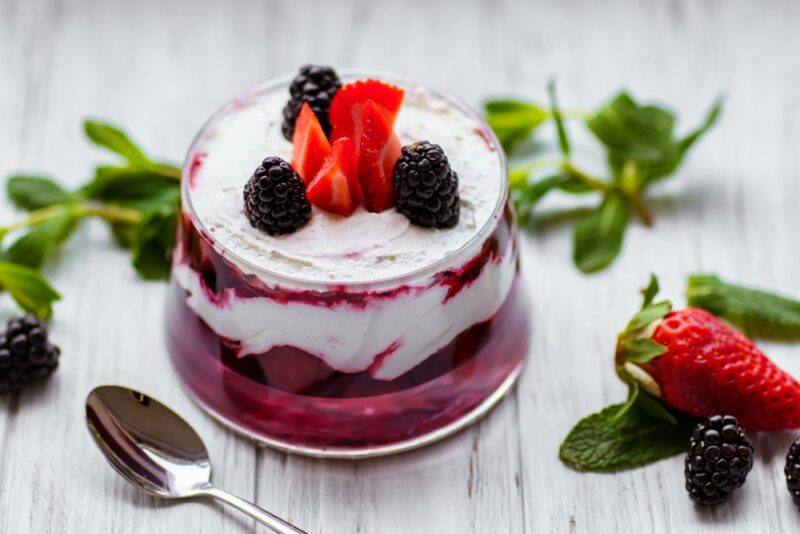 A glass jar containing a cheesecake parfait with berries on top, next to a spoon and some leaves