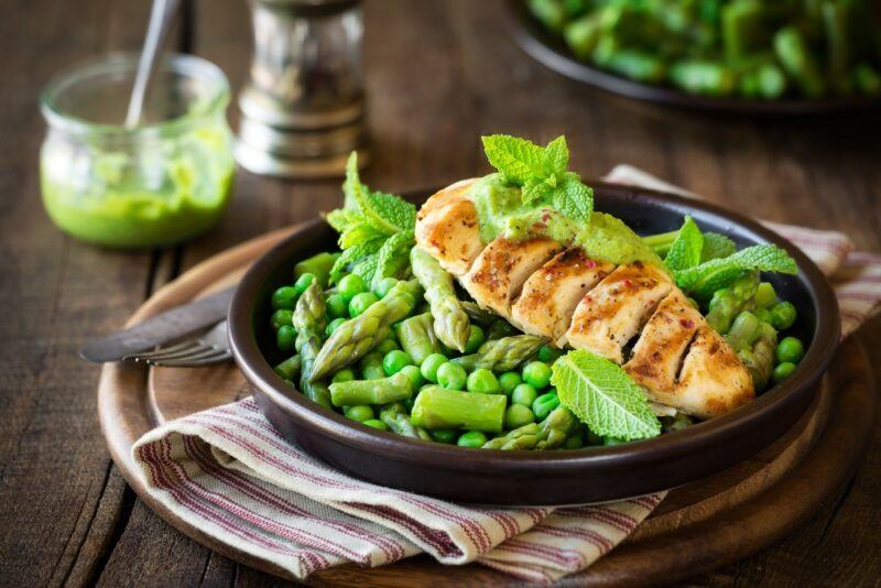 A black plate at a dinner table, with peas, asparagus, and fresh chicken breast