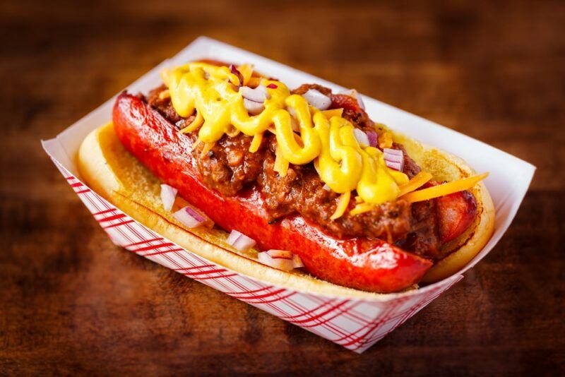 A paper container with a single chili dog, complete with onions, ground beef, a sausage, and plenty of mustard