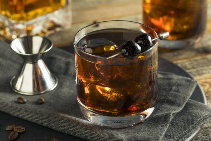 A chocolate peanut butter old fashioned in a cocktail glass, garnished by two olives, next to a cocktail jigger, a bottle of spirits, and a second cocktail in the background