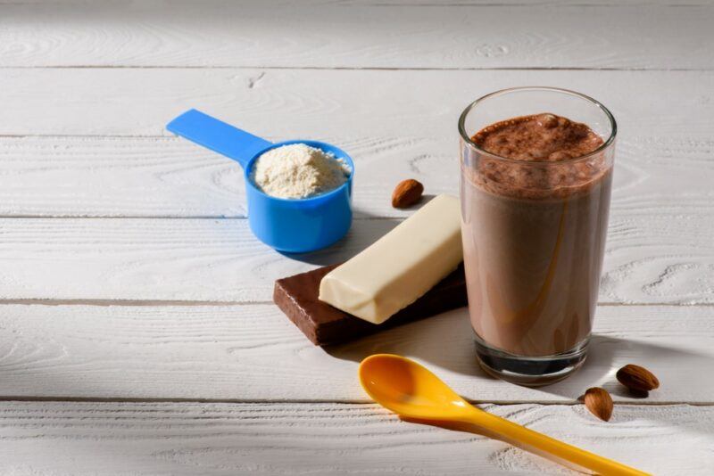 A chocolate protein shake in a glass, next to a protein bar and a scoop of protein powder