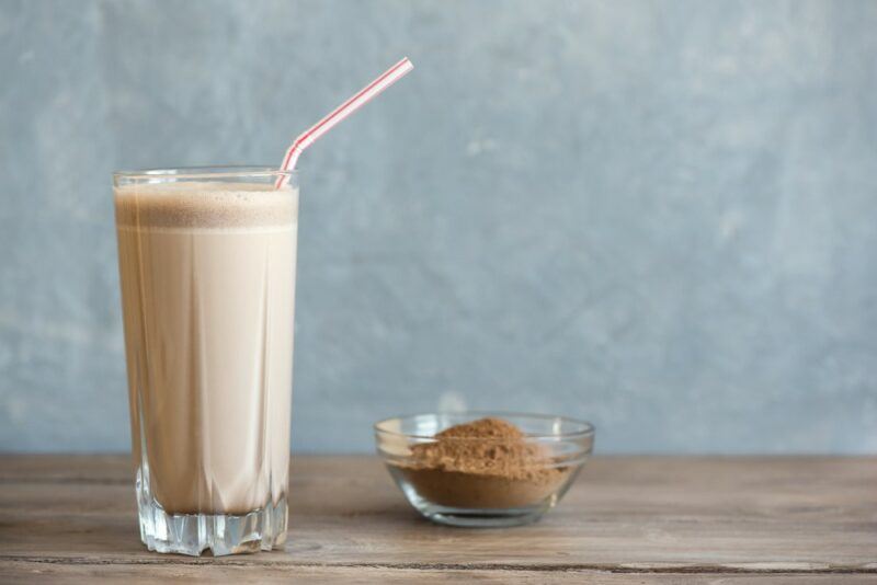 A glass of a light chocolate protein shake, next to a small glass dish of the powder