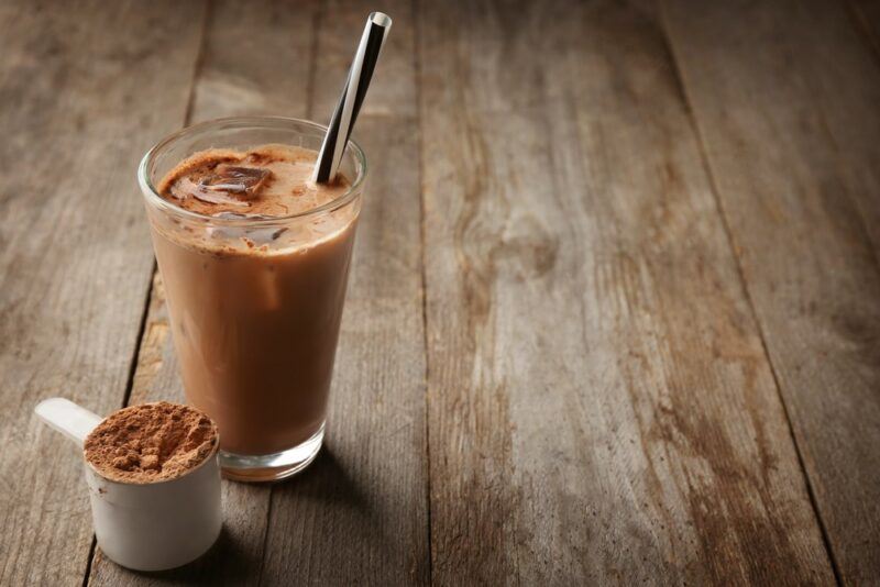 A wooden table with a glass of chocolate protein shake and a scoop of chocolate protein powder