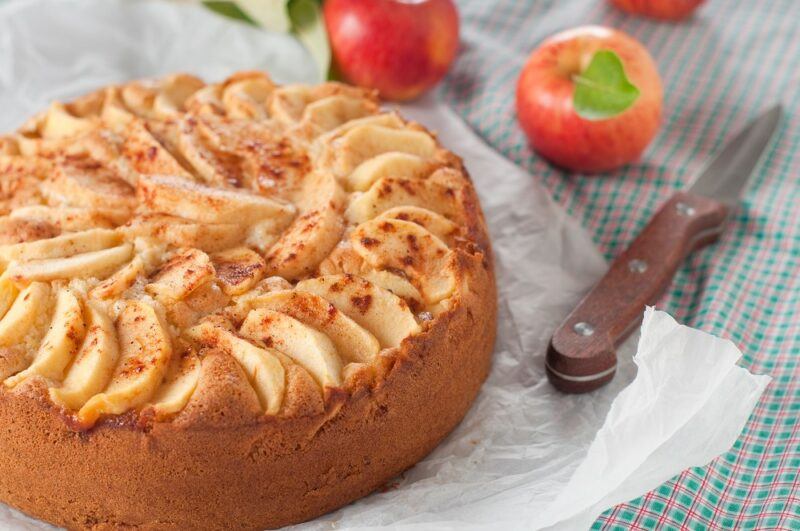 A glass plate with a cinnamon and apple cake next to a knife, with a few apples in the background