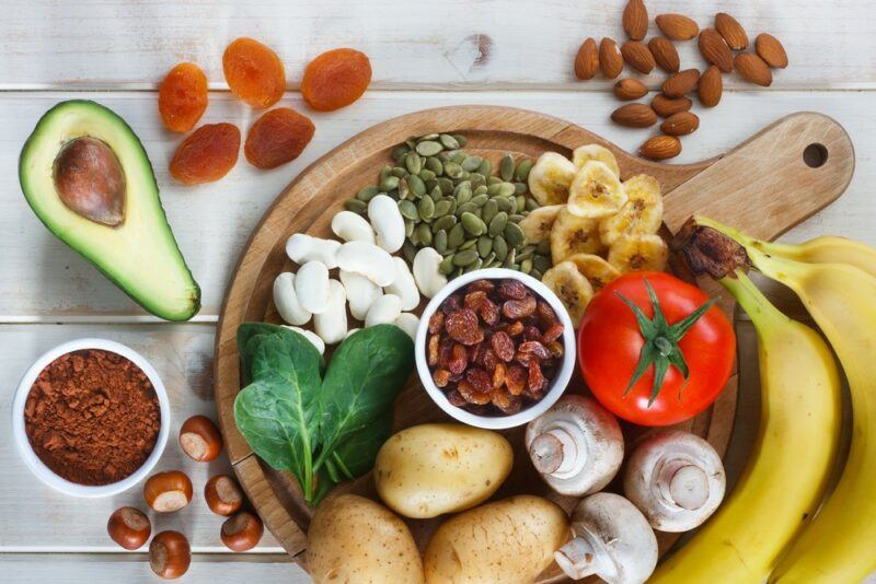 A circular wooden board with a selection of potassium rich foods, like legumes, dried bananas, fresh bananas, potatoes, and mushrooms, with a few other fresh ingredients on the table surrounding the board