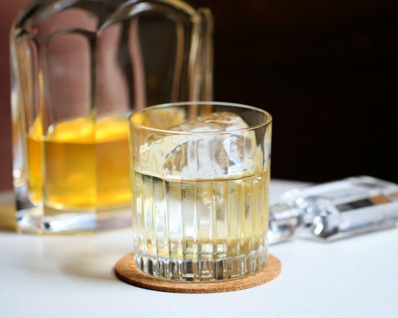 A glass of a clarified milk punch with ice, in front of a bottle of spirit