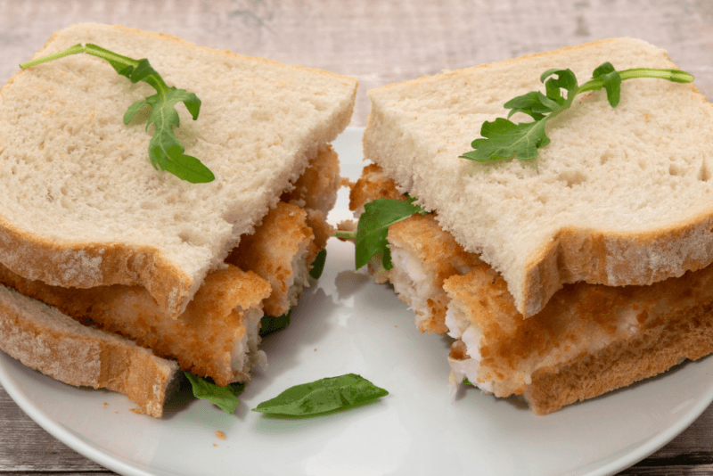 A fresh fish finger sandwich on a white plate. The sandwich has been cut in half, so you can see the interior of the fish fingers.