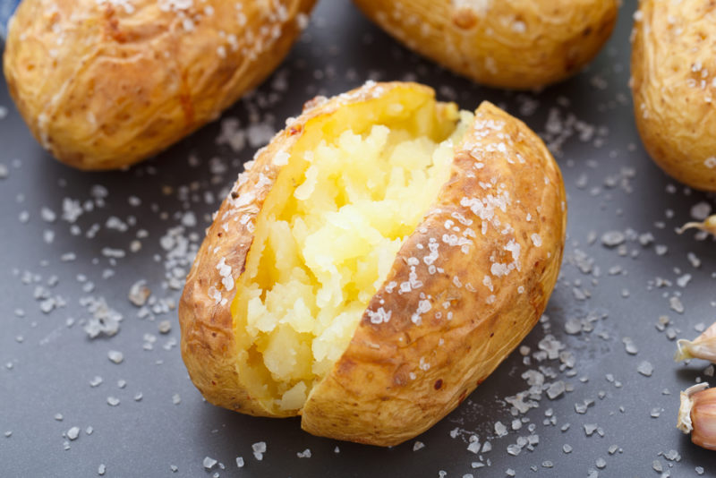 A single baked potato that has been cut open and sprinked with salt, with a few whole baked potatoes in the background